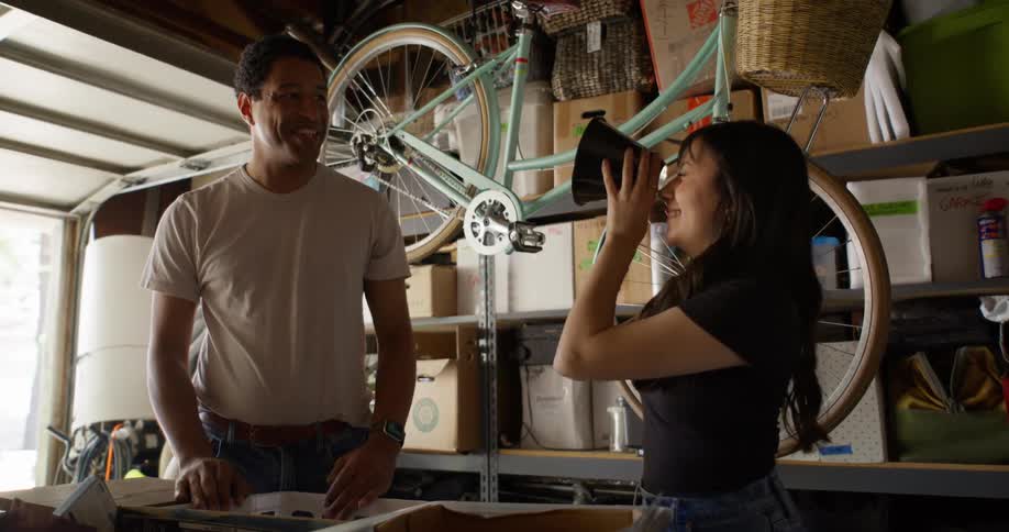 A woman takes a cylindrical object out of a box and places it on her face to observe a man who is next to her and they laugh