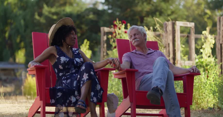 A couple are sitting on red adirondack chairs in a garden holding hands and enjoying a sunny day