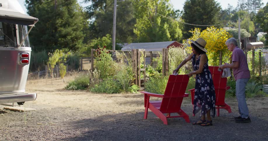 Two people are arranging red adirondack chairs outside near a silver trailer with a garden and trees in the background