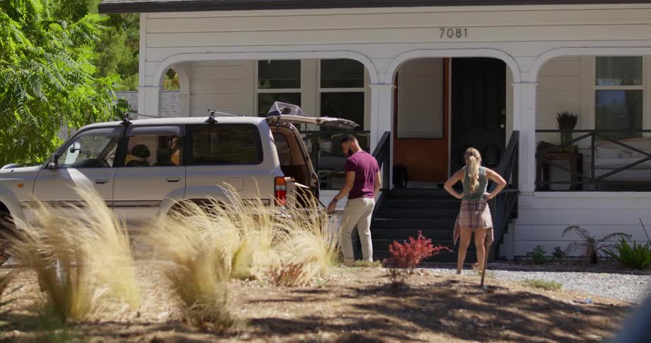 Man opening the trunk and a woman next to him seeing a house next door