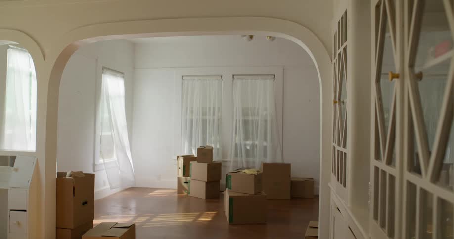 A large empty living room with white walls and wooden floors is filled with cardboard boxes suggesting a recent move