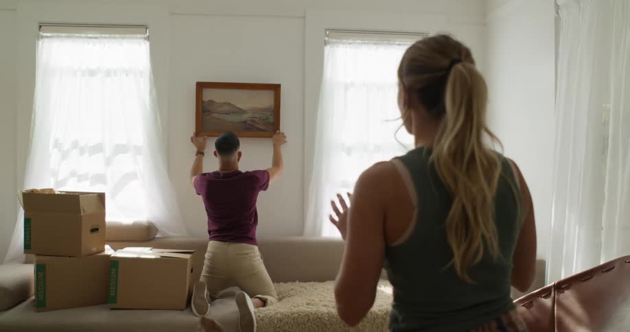 A woman gives her partner instructions on how to align a painting above a couch