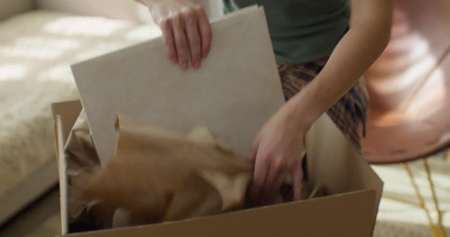 A person unpacks a box filled with brown packing paper they remove a large white photo album from the box they hold the album in their hands