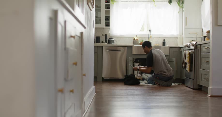 A man is fixing a kitchen sink in a welllit kitchen at home