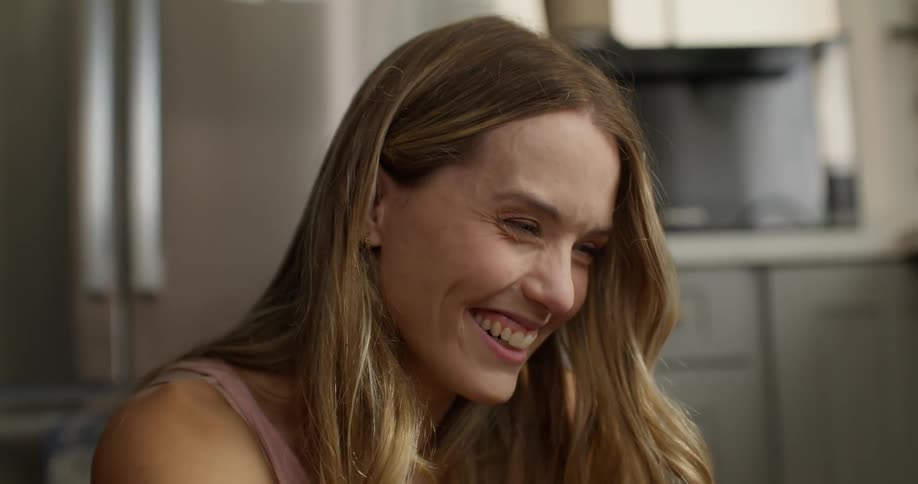 Blonde woman with a pink top bursts into laughter in the kitchen