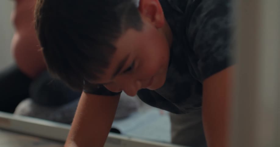 A young boy cleans the floor as his mother rests in the background