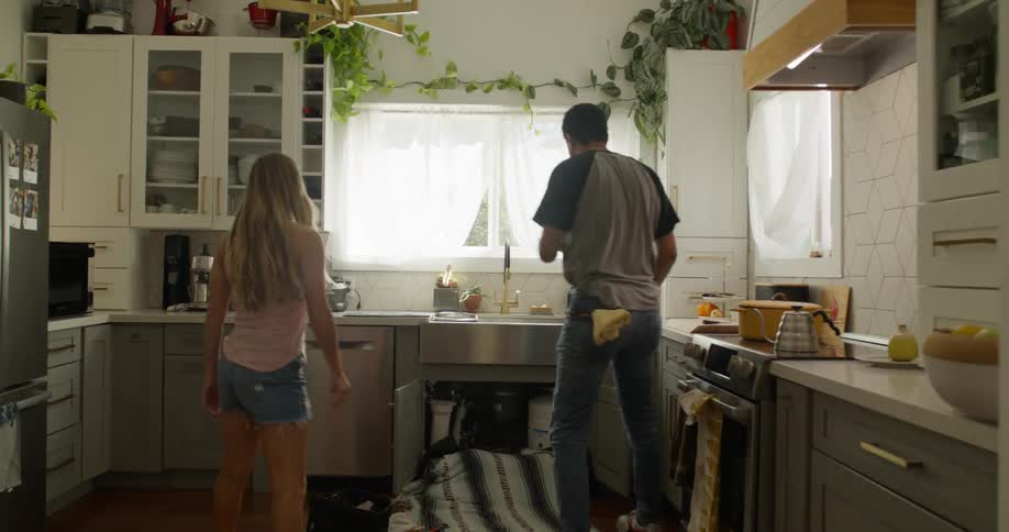 A woman is kneeling on the floor of a modern kitchen looking at a plumber fixing her sink