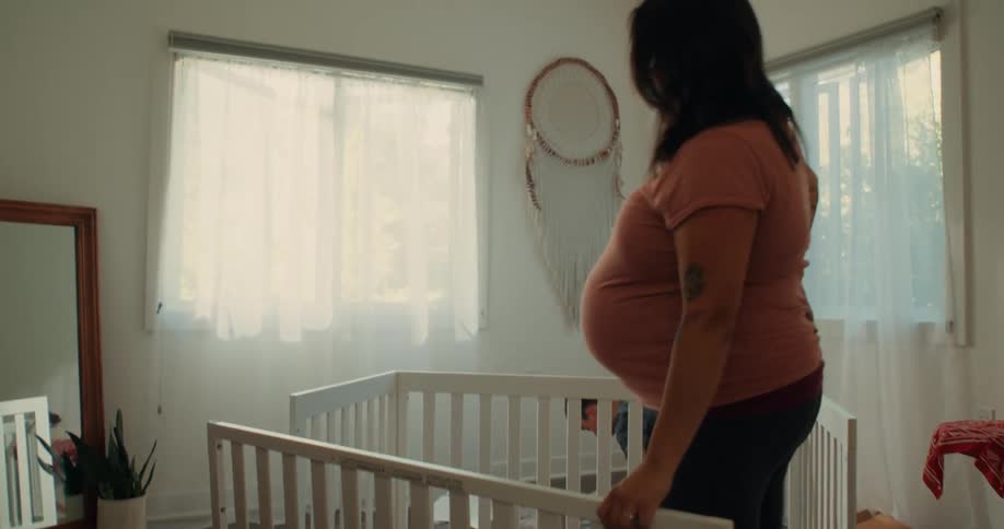 A pregnant mom with arm tattoos wearing a pink shirt helps her son assemble a crib in a room