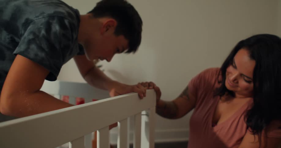 A son carefully uses a screwdriver to assemble a crib while his pregnant mom watches in the room