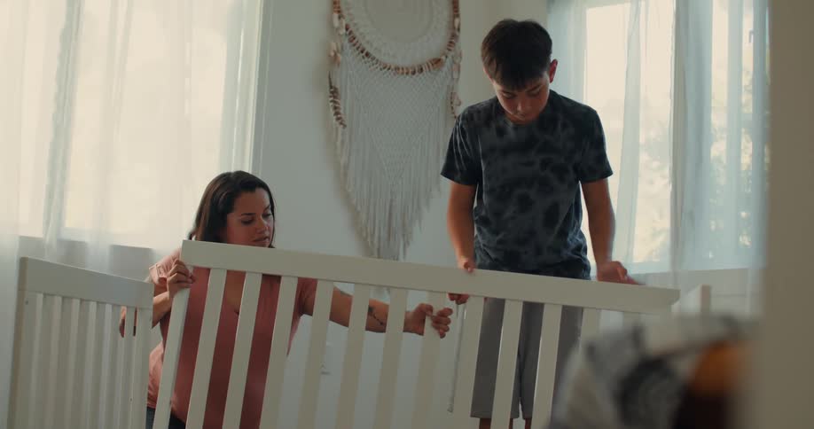 A pregnant mother and her son work together to assemble a crib in the room