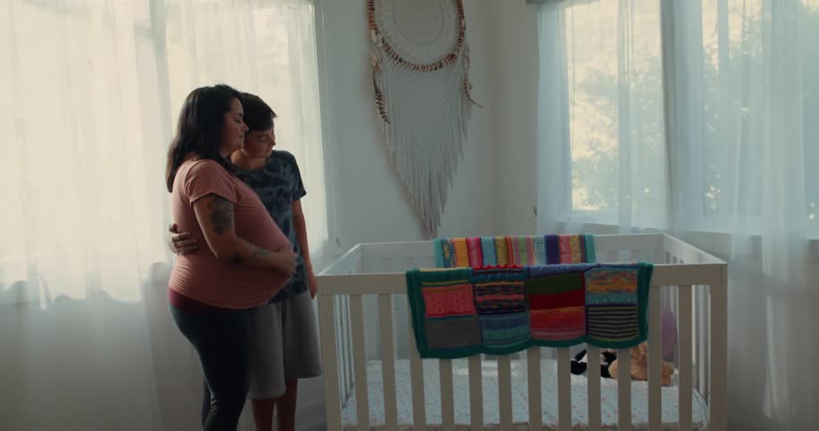 A pregnant mother and her son holding each other look down at a new crib set in the room