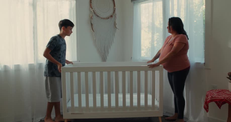 A teenage boy and a pregnant adult woman are carrying a white wooden crib to a corner of the room