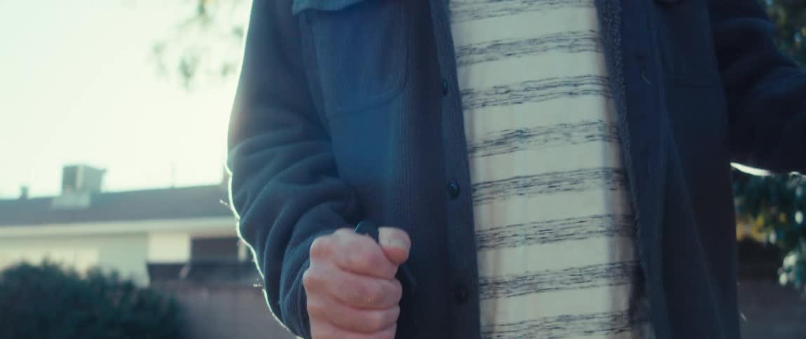 The sun shines brightly as a man in a striped shirt and navy blue jacket strides down the street trekking poles in hand