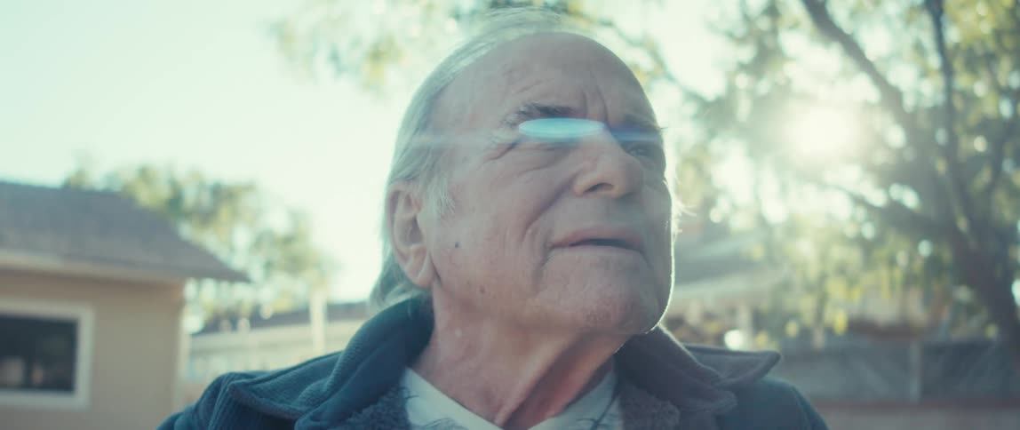 The face of an elderly man with gray hair who is looking up in an outdoor setting with the sunlight behind him