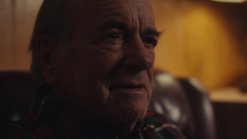 An elderly man sits in a dimly lit room his thoughtful expression and subtle smile conveying warmth and a sense of nostalgia