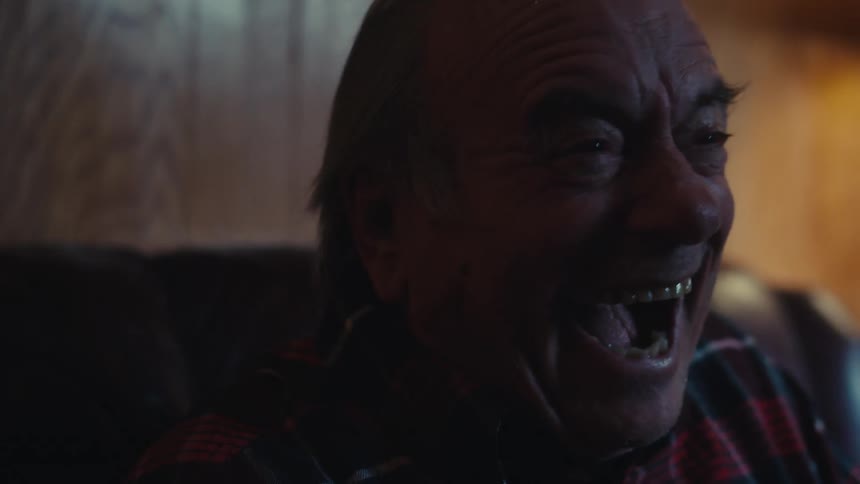 An old man in a red plaid shirt watches tv his expression turning from calm to frantic joy