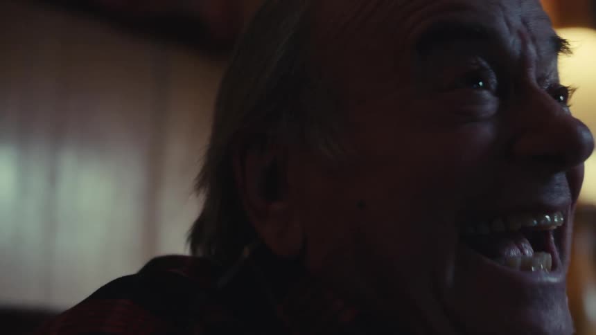 An elderly man sitting on a sofa looking forward is smiling and celebrating happily