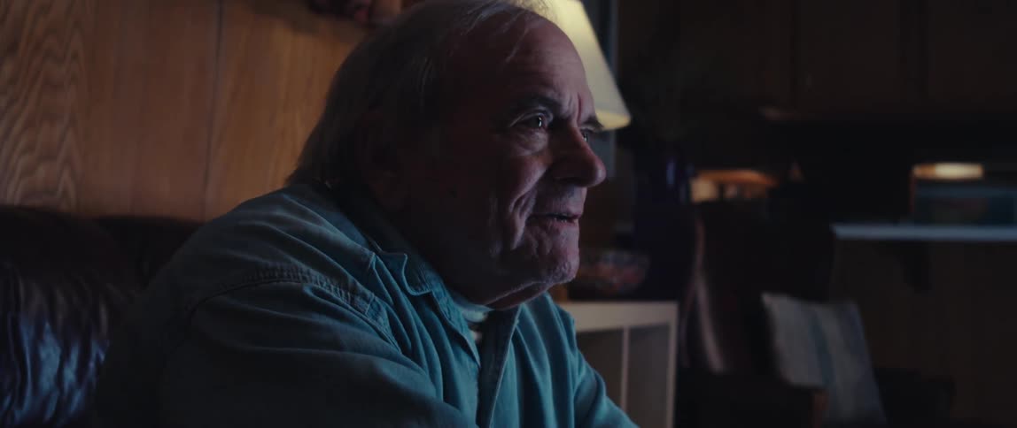 An elderly man in a denim jacket plays a video game in a dimly lit room determined to win