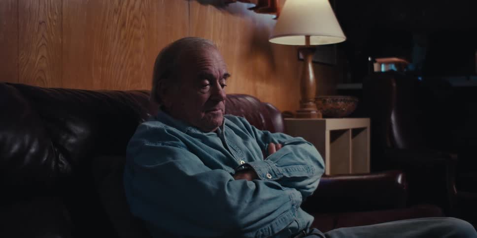 An elderly man wearing a denim jacket and with arms crossed sits on a brown leather couch in a dimly lit room