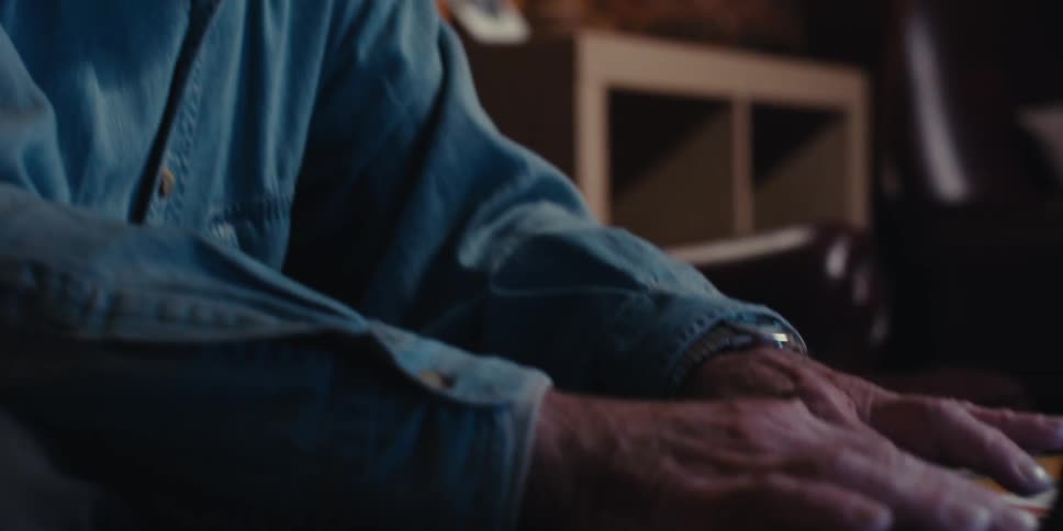 An elderly man wearing a denim shirt and glasses types on a laptop in the room