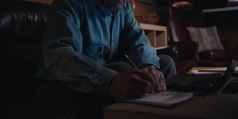 Elderly man sitting in front of a laptop and with a pen writing in a notebook