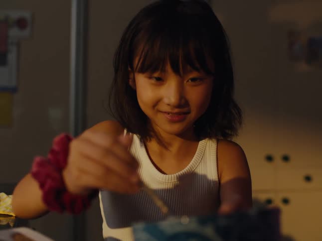 The face of a happy little girl who is stirring a spoon in a blue bowl and she is pushing an adults hand