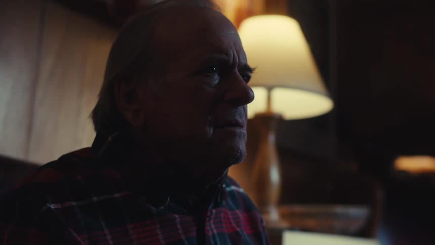 An older man in a plaid shirt sits on a couch and talks in a dimly lit room