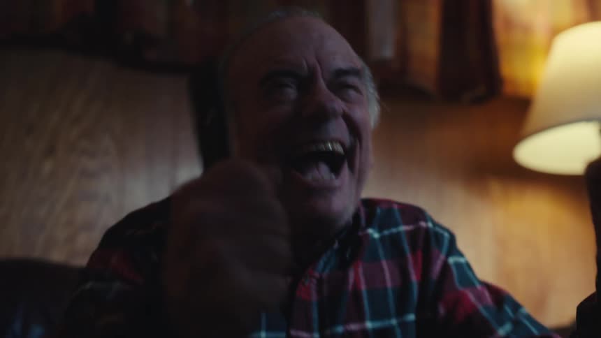 An elderly man wearing a red plaid shirt looks up and begins to laugh heartily while sitting in a dimly lit room