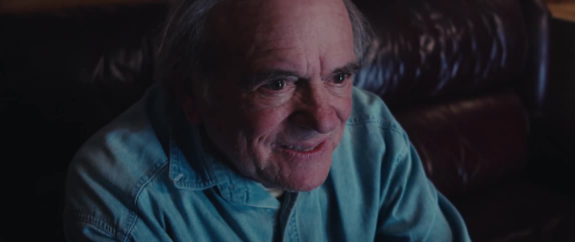 An elderly man sitting on a couch bursts into laughter while watching television