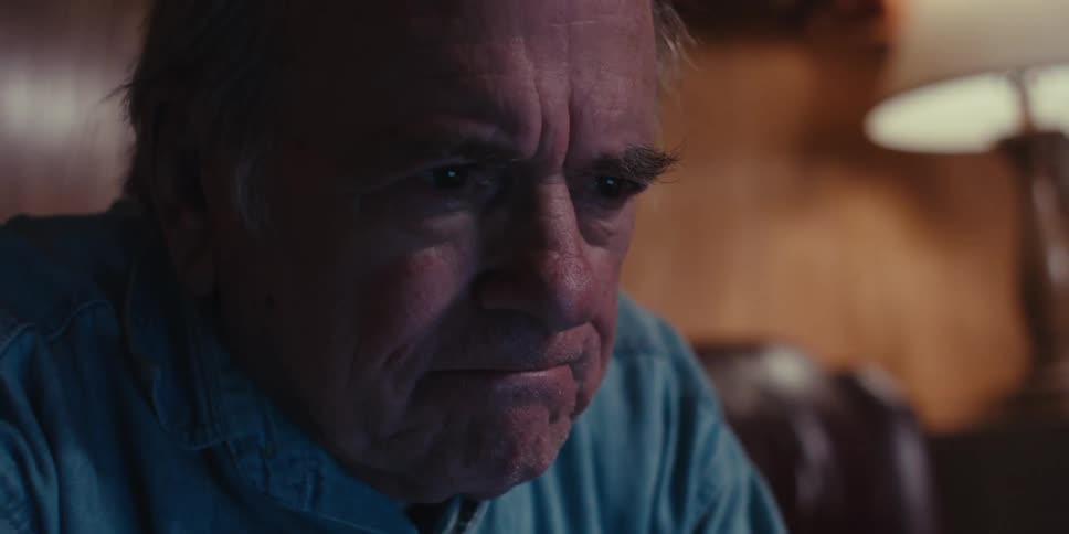 An elderly man in a blue shirt sits at a laptop in a dimly lit room his eyes filled with sadness