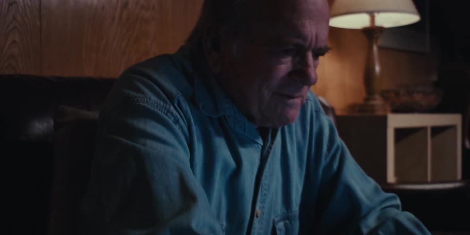 A pensive elderly man wearing a blue shirt sits on the couch and uses a laptop in a dimly lit room