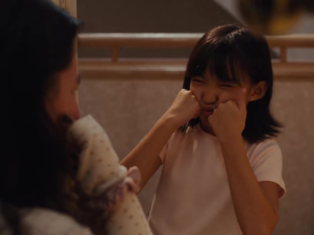 A little girl is putting her fingers on her cheek and making faces at a person in front of her