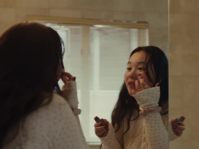 A young woman puts on lipstick while looking in a bathroom mirror