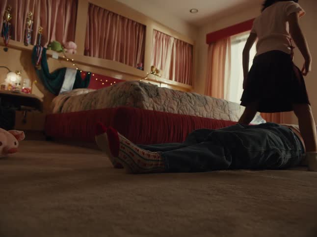A little girl jumping over an adult who is lying on the floor of a room with a bed next to it