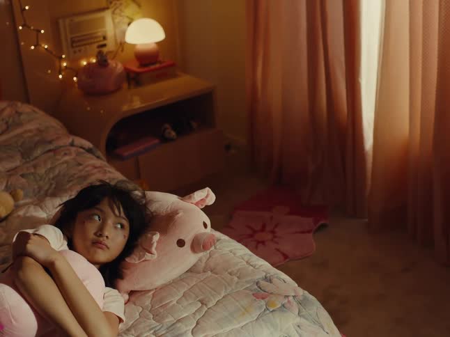 A girl is lying in bed with plush pigs beside her near a window covered with curtains