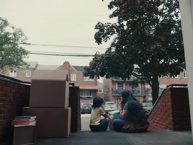 Two people are sitting on the stoop of a building surrounded by boxes looking out at a residential street and talking