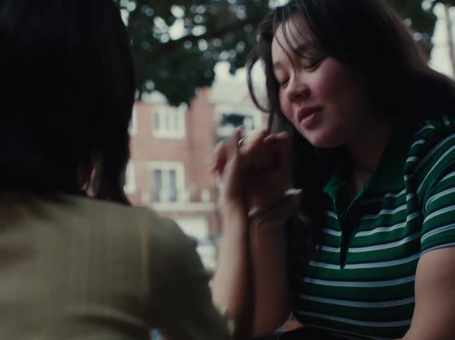 One young person rests their head on their hand and listens attentively while another young person whose face is not visible speaks to them outdoors