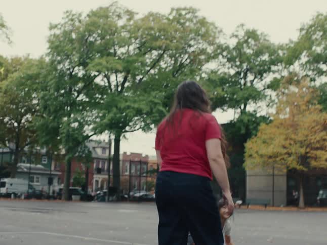 Two people one a younger girl are playing with a blue ball on an outdoor basketball court with trees and buildings in the background