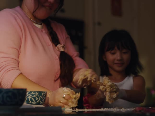 A person in a pink cardigan and a child are working together to knead dough on a kitchen counter with bowls and other baking items around them