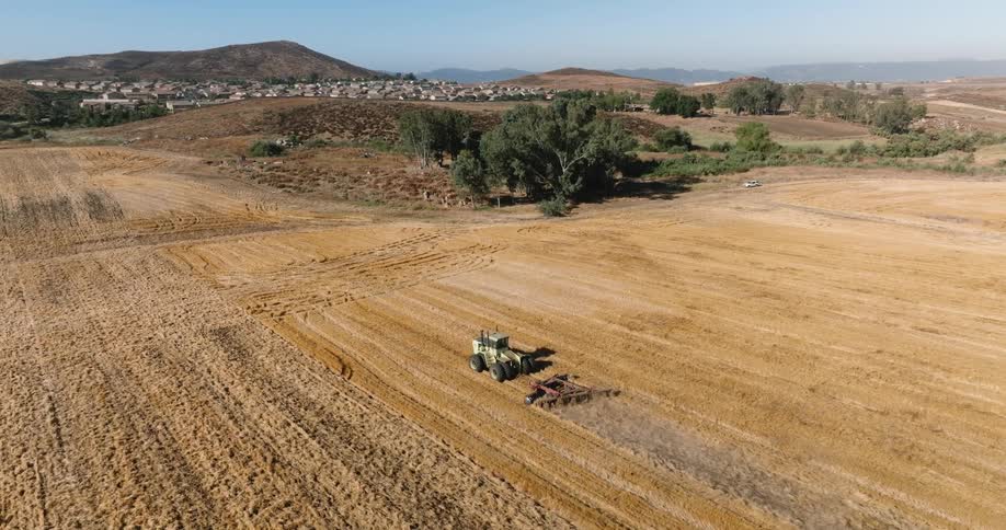 A tractor tills a vast golden field under a clear blue sky with rolling hills and a distant town in the background