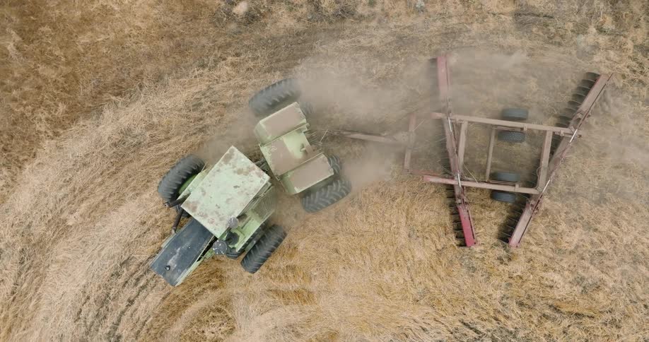 Tractor pulls a tilling attachment across a fenced field during the day