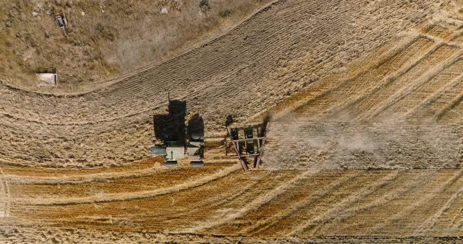 An aerial view of a tractor plowing a large dry agricultural field leaving visible curved lines in its wake as it tills the soil