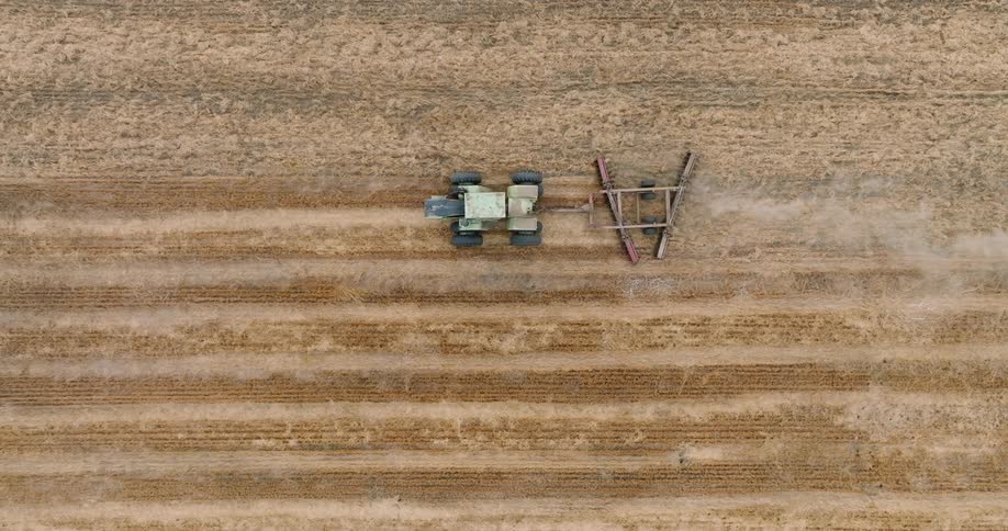 A tractor with an attached farming implement drives in a straight line across a harvested field
