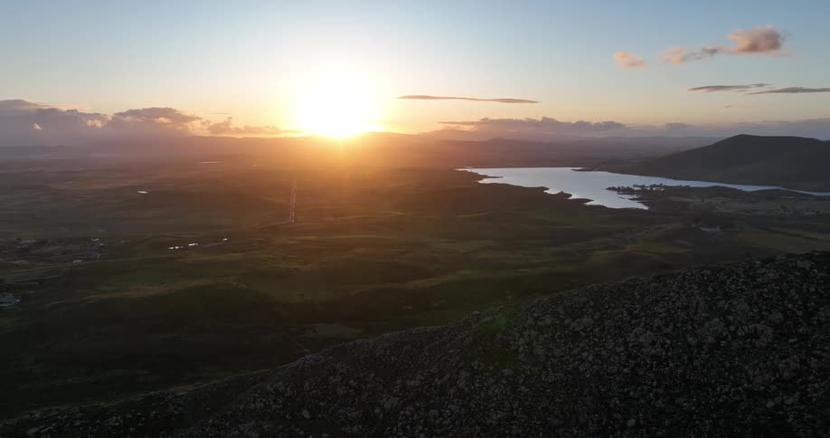 An aerial wide shot shows the sun setting over a vast rocky landscape with a lake and mountains in the distance