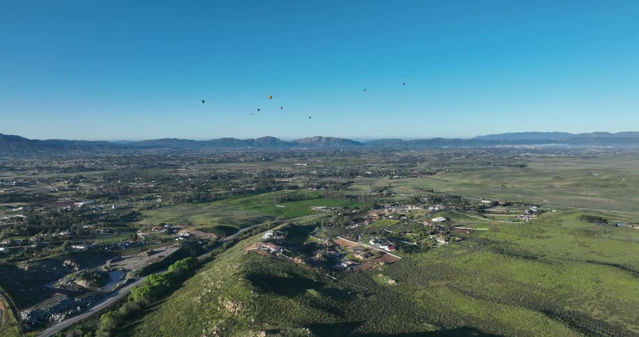 A wide aerial shot shows a landscape with hills and a city with several hot air balloons floating in the clear blue sky