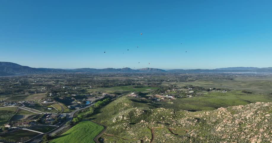 Aerial view of a scenic landscape and a sprawling valley with scattered houses and roads while several hot air balloons are visible in the clear blue sky