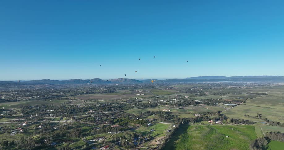 A large field with green vegetation and small green hills and some hot air balloons flying over in the background