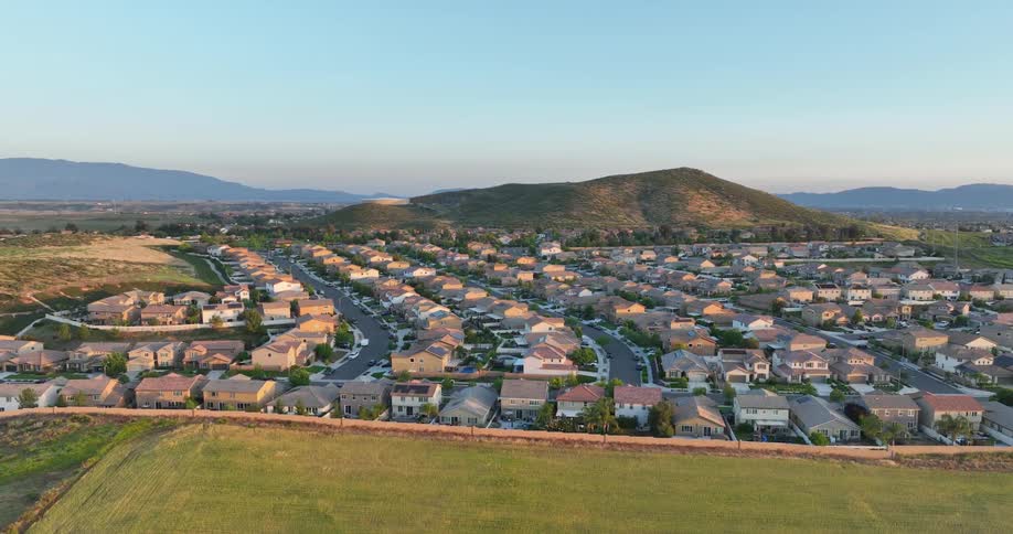 A sprawling suburban neighborhood nestles against a backdrop of rolling hills bathed in the warm glow of the setting sun