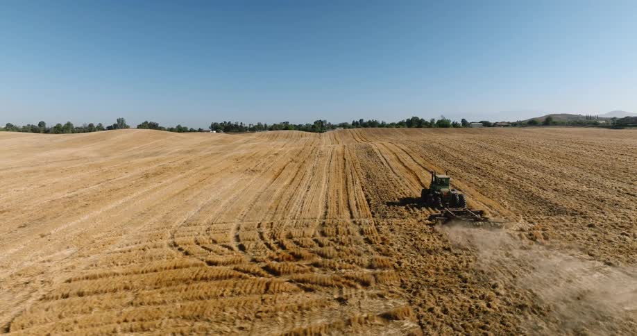An agricultural tractor with a combine harvester behind passing through a farm field with beige soil