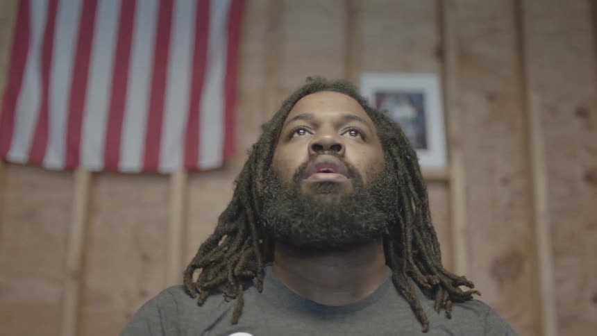 A man with a beard is looking up with a wall and an american flag in the background